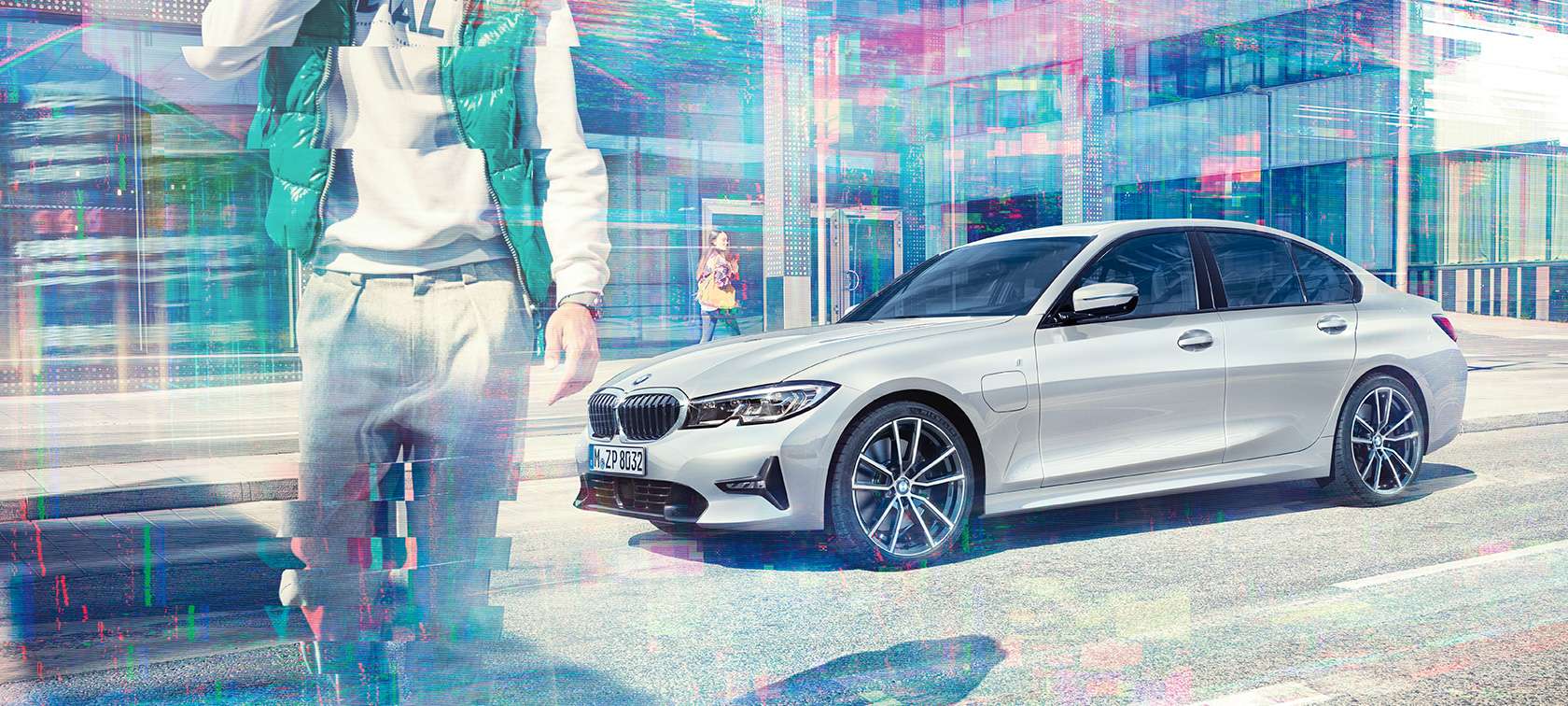 BMW 3 Series Saloon, side shot BMW 330e. Three-quarter side shot of the standing BMW 330e Saloon in front of an urban glass building with walking passengers.