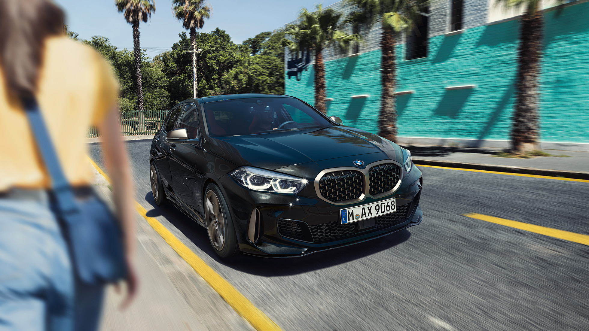 BMW M135i xDrive, three-quarter front view. BMW M135i xDrive in Black Sapphire metallic, three-quarter front view driving, with woman in foreground.