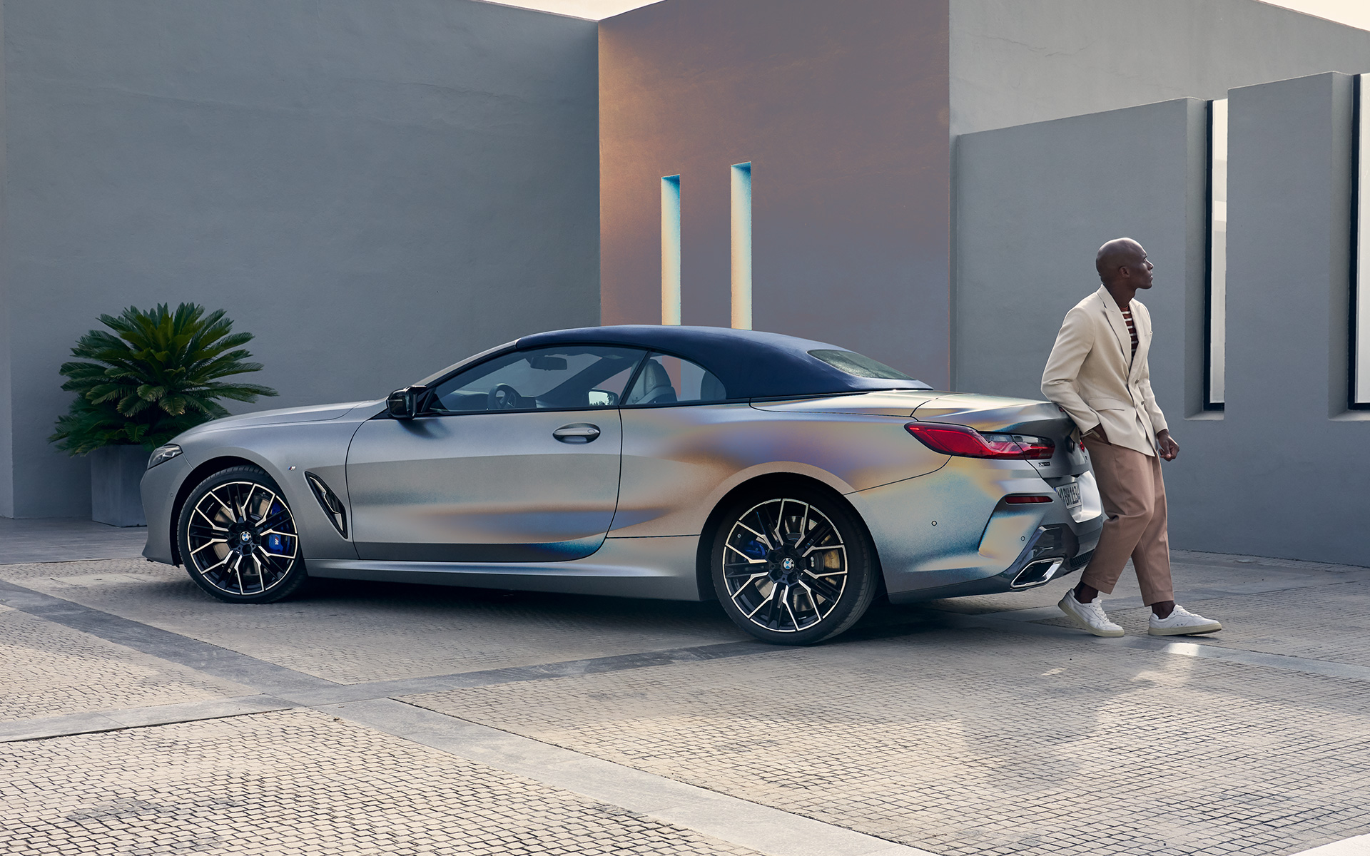 Three-quarter rear view BMW M850i xDrive Convertible G14 LCI Facelift 2022 BMW M850i xDrive Convertible G14 LCI Facelift 2022 BMW Individual Frozen Pure Grey metallic three-quarter rear view with male model leaning against vehicle