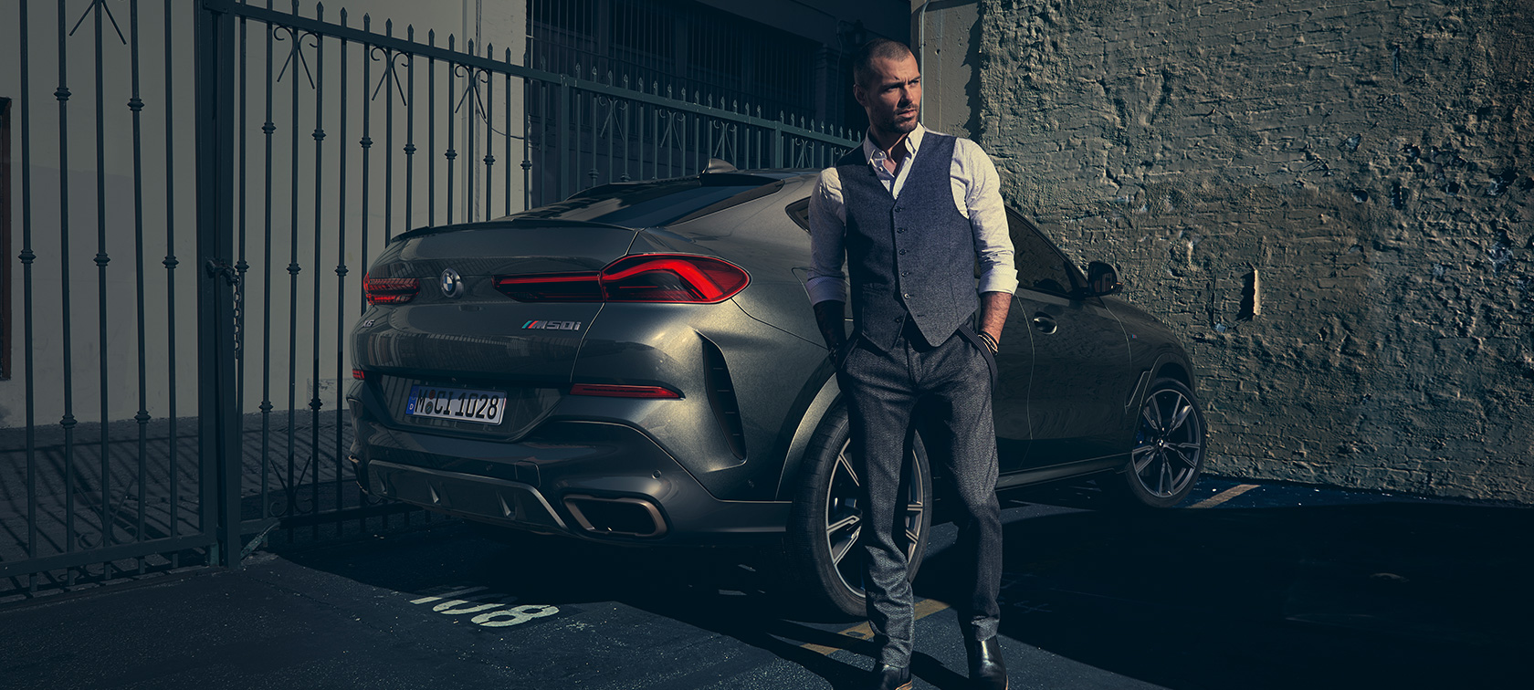 BMW X6 M50i in three-quarter rear view. Man standing next to BMW X6 M50i SUV in three-quarter rear view.