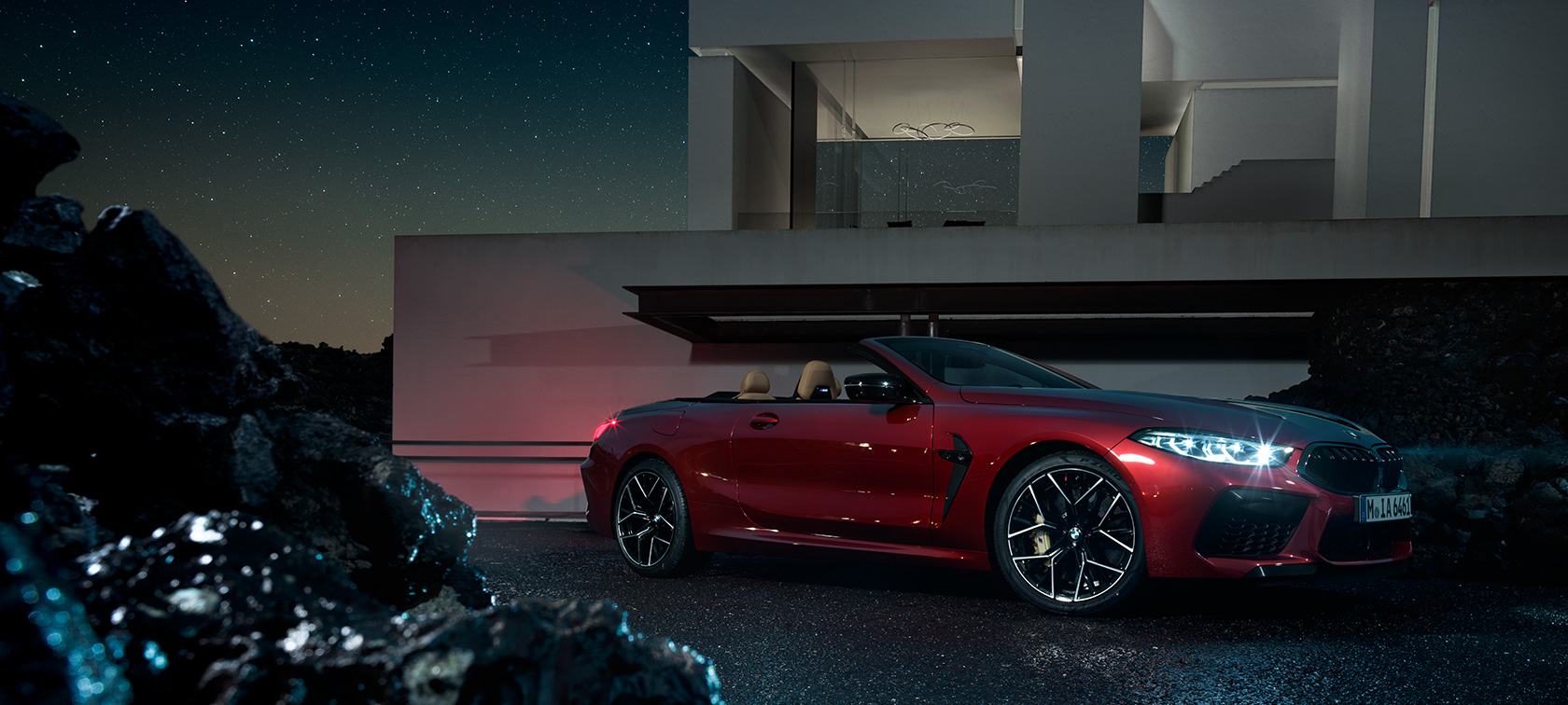 BMW M8 Competition Convertible, three-quarter side view. BMW M8 Competition Convertible in Motegi Red metallic, three-quarter side view, standing at night in front of villa.
